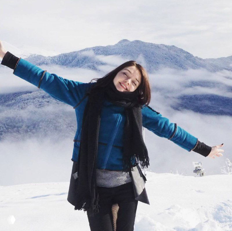 Автор в зимней куртке улыбается и раскинула руки на фоне заснеженных гор; на фото видна снежная панорама и ясное небо