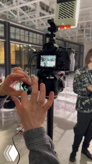 Крупный план видеокамеры на штативе и руки оператора во время съёмки в помещении; на заднем плане люди и интерьер мероприятия.