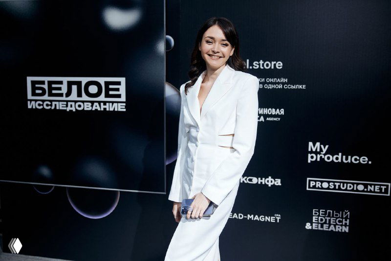 Автор в белом костюме стоит на фоне баннера мероприятия с надписями «БЕЛОЕ ИССЛЕДОВАНИЕ» и логотипами спонсоров, улыбается в камеру