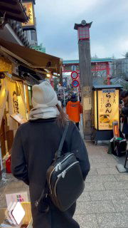 Человек в тёплой одежде идёт по узкой туристической улице в Японии, видны вывески, лавки и прохожие — уличная туристическая сцена.