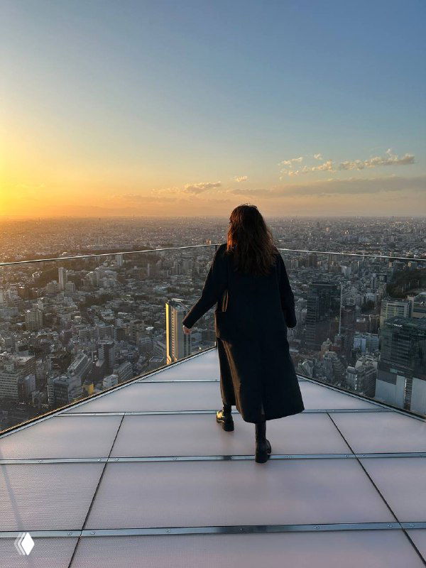 Женщина в пальто стоит на крыше высотки и смотрит на город на закате — образ фокуса, амбиций и масштабных целей клиентов.