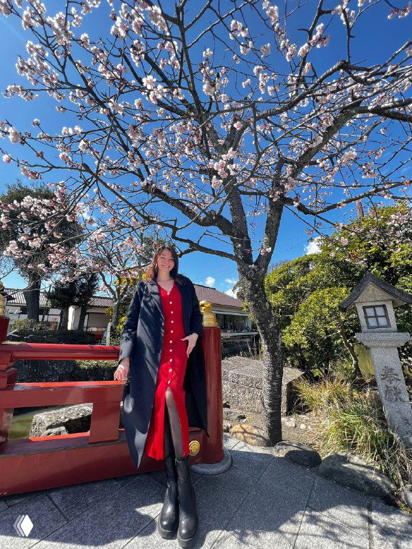 Женщина в красном платье и пальто стоит у красного перила рядом с цветущим деревом и каменными фонарями на ярком небе.