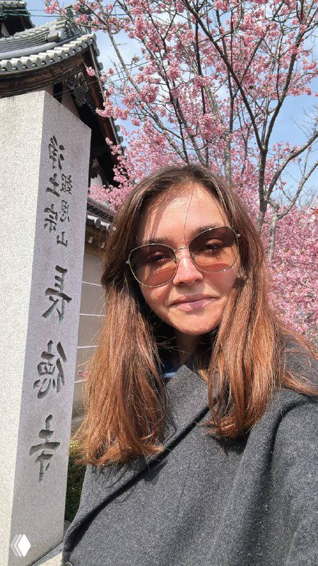 Портрет женщины в очках на фоне цветущей сакуры и каменной колонны с вертикальными надписями, уличный кадр в сезон цветения.