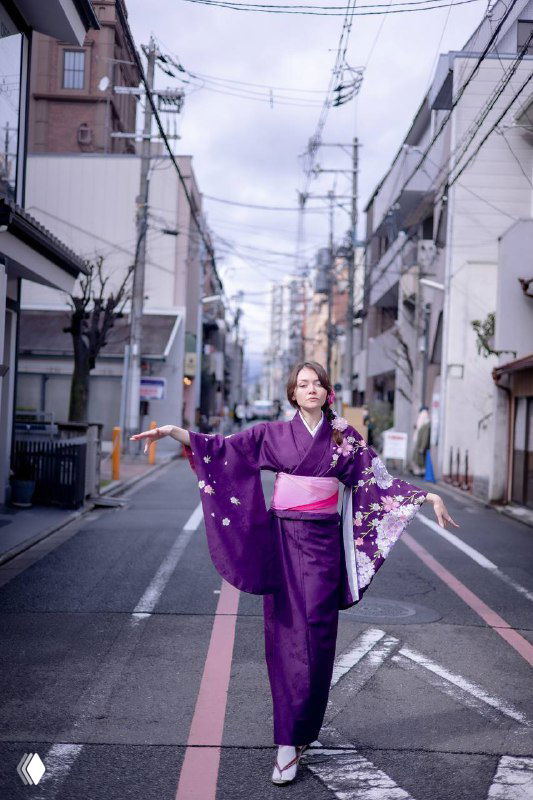 Женщина в фиолетовом кимоно стоит посреди узкой улицы японского города, руки разведены в стороны, видны дома, дорожная разметка и электрические провода.