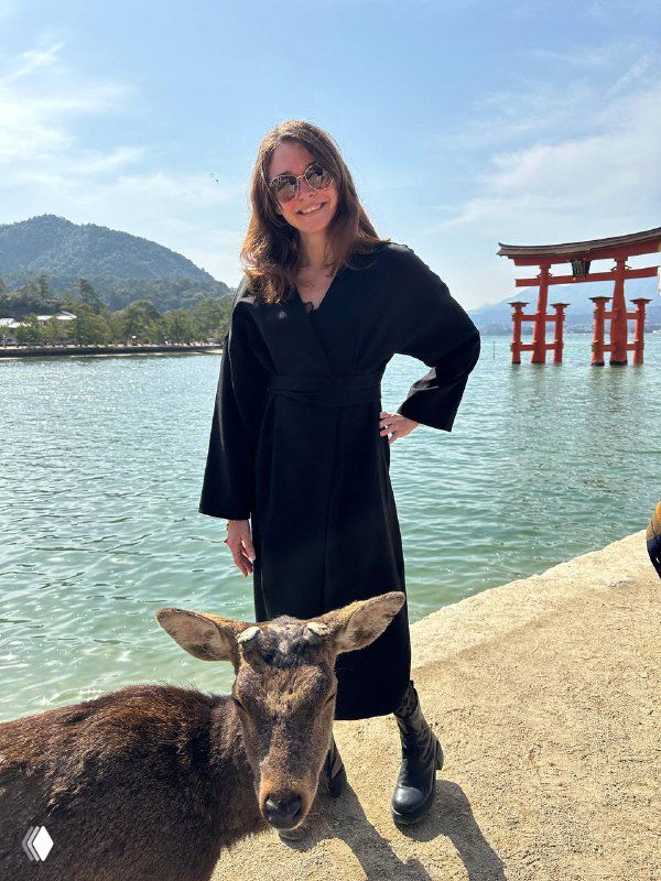Женщина в чёрном плаще стоит у кромки воды и улыбается; рядом доверчивый олень, на заднем плане видна тории, озеро и горы.
