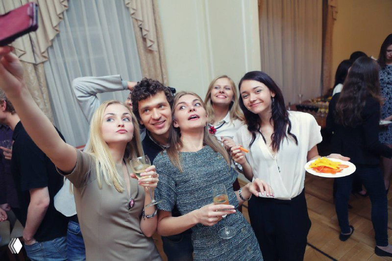 Группа людей на корпоративе: несколько участниц делают селфи, держат бокалы и тарелку с закусками; фотография из подборов персонала.