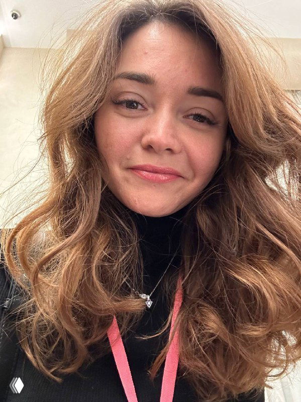 Портрет улыбающейся женщины в интерьере, крупный план лица, естественная причёска и мягкое дневное освещение, нейтральный фон.