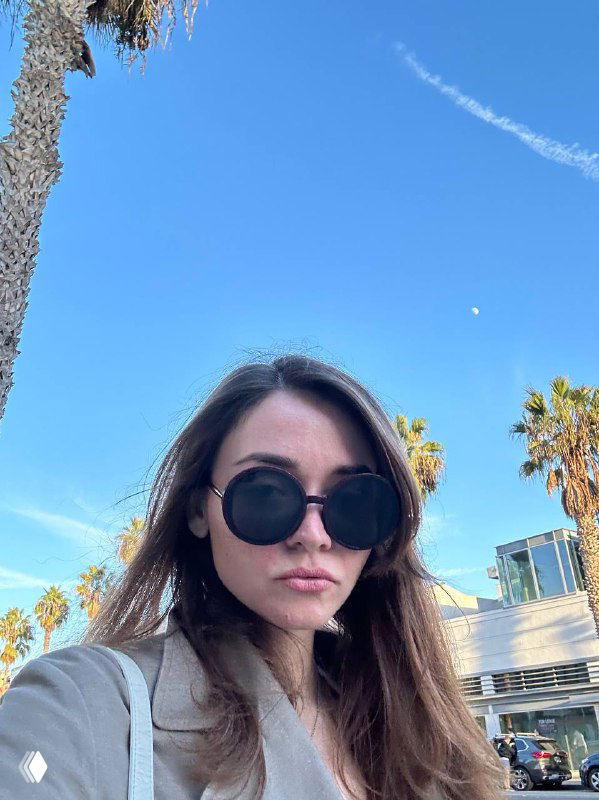 Портрет женщины-автора блога в деловом образе: тёмные очки, распущенные волосы, пальмы и голубое небо, летний городской фон.