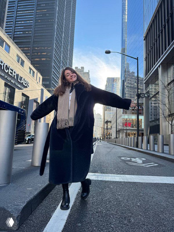 Женщина в тёмном пальто и шарфе идёт по городской улице с небоскрёбами; деловой городской фон, стильная прогулка.