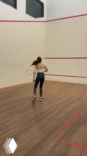 На фото женщина с ракеткой стоит на корте для паделя, снята сзади: момент тренировки, движение к мячу на деревянном полу в закрытом зале.