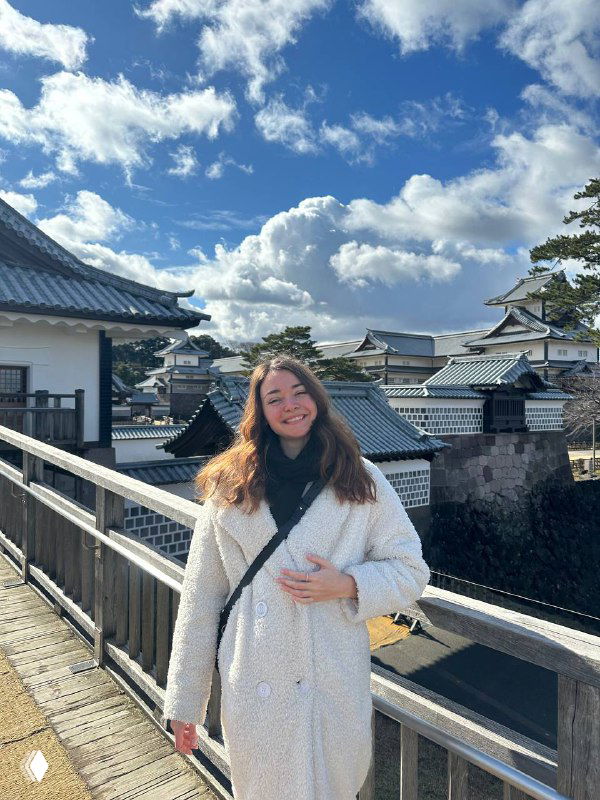 Женщина в белой шубе стоит на деревянном мосту на фоне традиционных крыш и голубого неба, улыбается в кадр — иллюстрация спокойствия и ресурсного состояния.