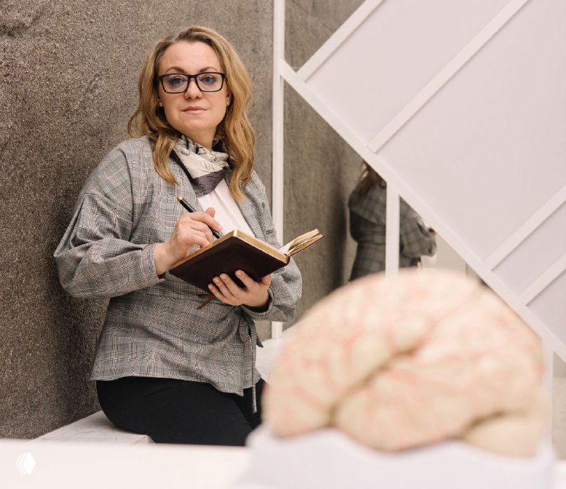 Нейропсихолог держит блокнот и ручку в кабинете; на переднем плане макет мозга — символ выгорания и синдрома самозванца.