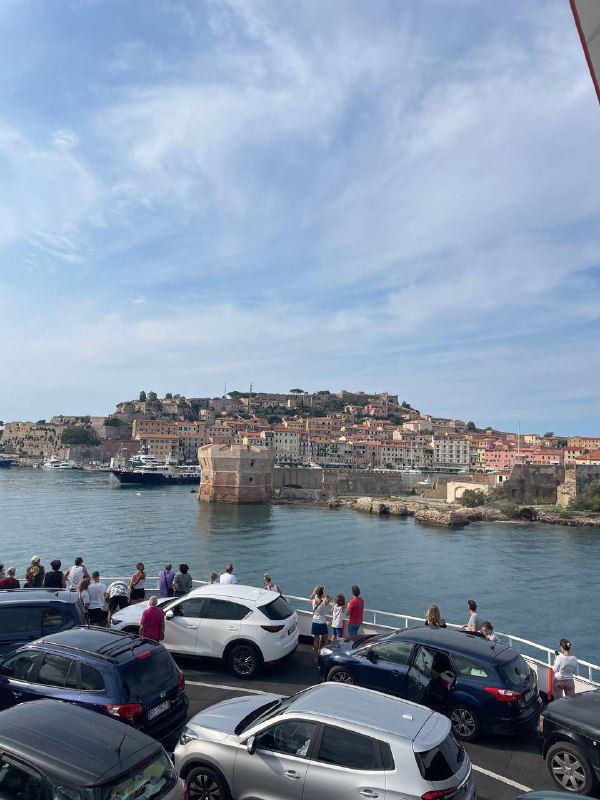 Порт и набережная острова Эльба: машины, люди у воды и круизные лодки на фоне прибрежного холма с домами — вид с причала на город.