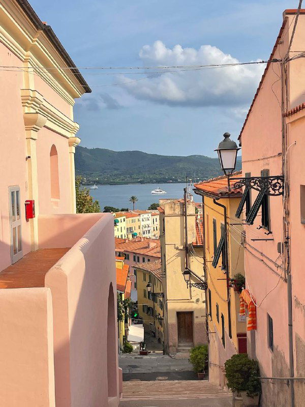 Узкая улочка с видом на море, розовые фасады домов и уличные фонари — тихая живописная сцена одного из рыбацких поселков Эльбы.