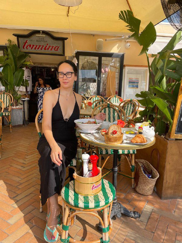 Кафе на улице острова Эльба: столик с едой и напитками, женщина в чёрном платье сидит рядом — сцена уютного обеда у моря.