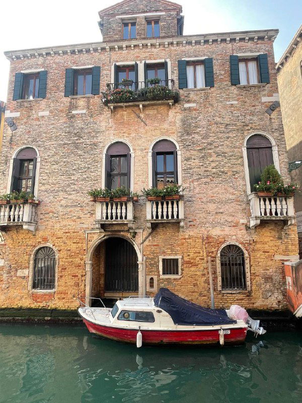 Кирпичное палаццо у канала с украшенными окнами и пришвартованной небольшой лодкой — типичная венецианская сцена у воды.