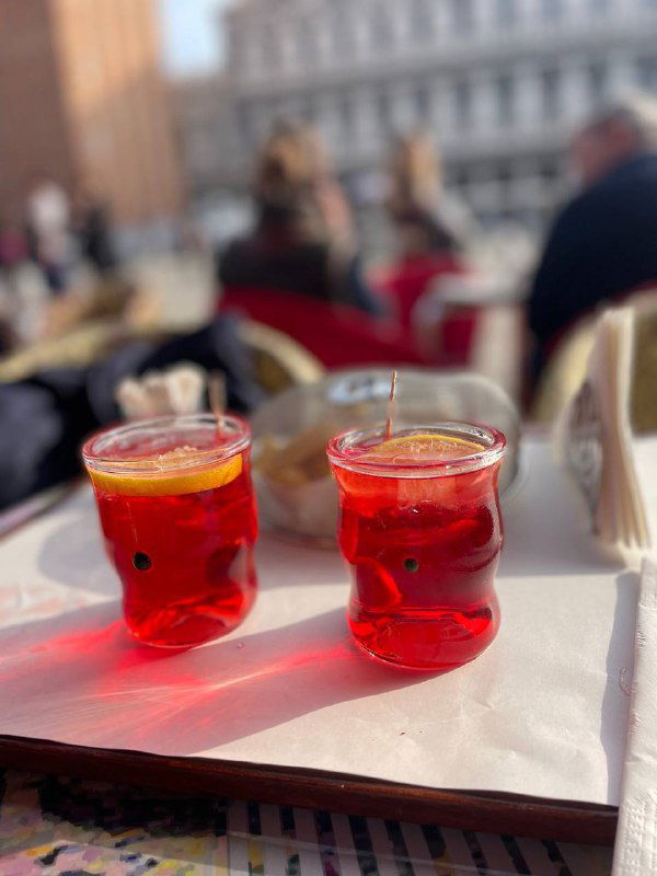 Крупный план двух бокалов спритца на столе, яркие апельсиновые оттенки напитка и размытие кафе на заднем плане в солнечном свете.