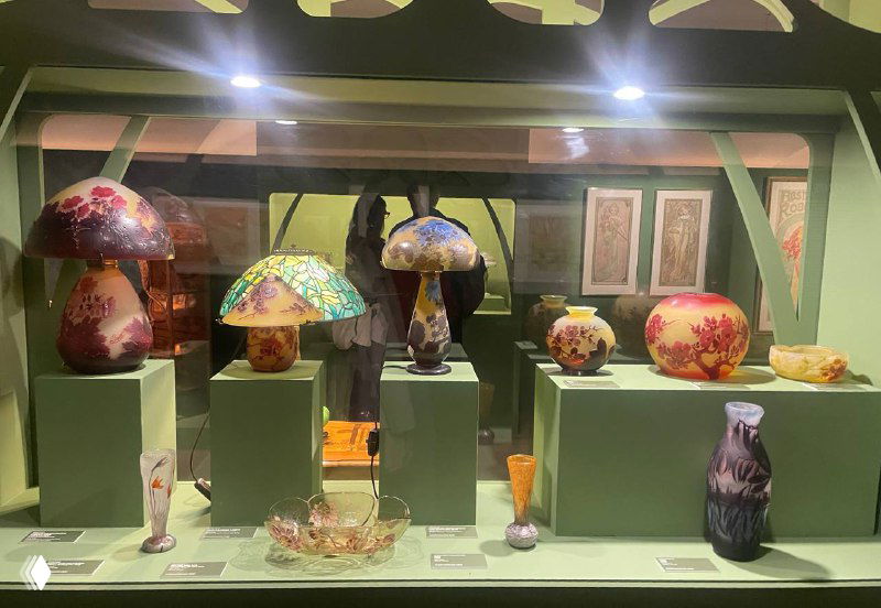 Витрина с коллекцией декоративных ламп и стеклянных ваз в музее: витражные плафоны в стиле ар‑нуво, размещённые на подиумах с зелёным фоном.