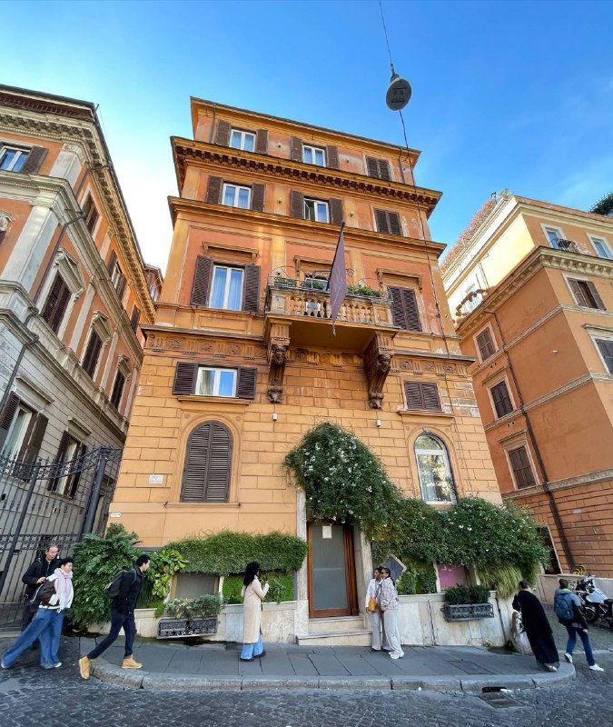 Оранжевый римский дом с балконами и зеленью у входа, прохожие и туристы у дверей — типичный городской фасад в Риме.