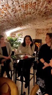 Трое женщин за высоким столом в книжном магазине Книгаголик; кирпичный свод, растение на заднем плане, атмосфера камерного вечера.