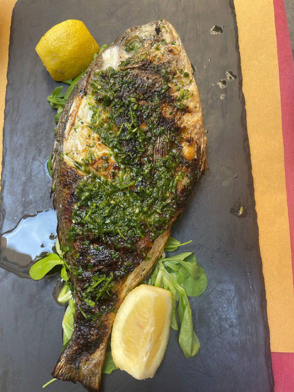 Жареная рыба на темной сланцевой тарелке с дольками лимона и зеленью, украшенная соусом песто — локальная кулинарная подача.