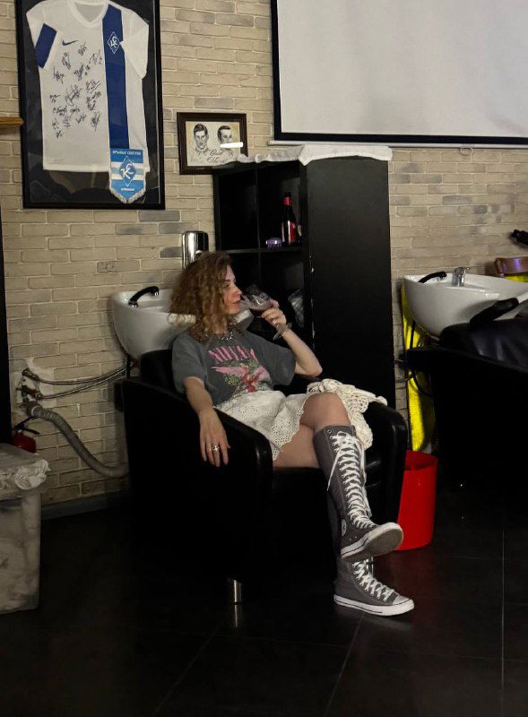 Гостья сидит в кресле для мытья головы в интерьере барбершопа Otto, на стене висит спортивная футболка и видна бытовая зона заведения.