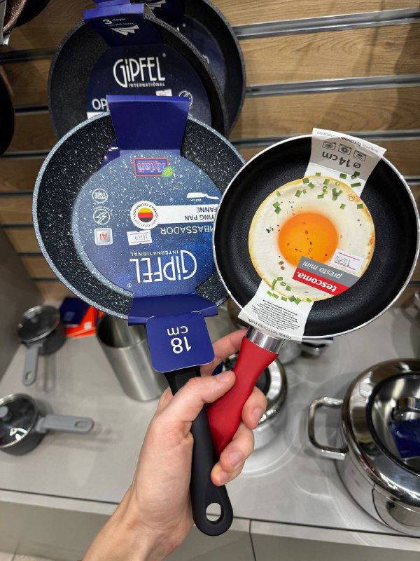 Рука держит две маленькие сковороды на витрине; одна с красной ручкой, видны бирки производителя и упаковка на полке магазина.