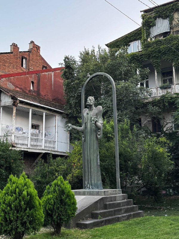 Скульптура женщины в небольшом садике на фоне старинных домов, поросших плющом; тихий дворик и зелёная растительность в центре города Тбилиси.