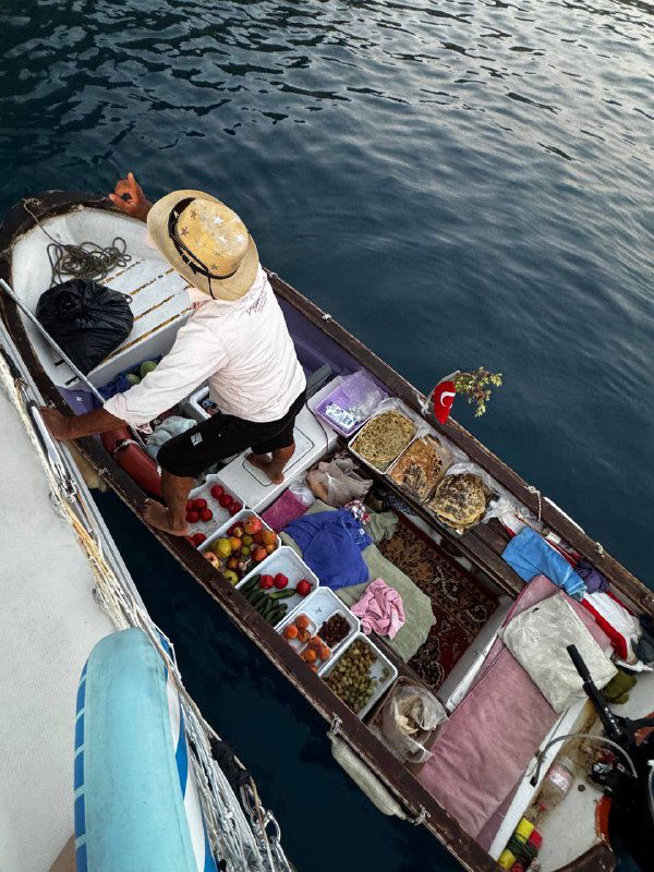 Ещё одна лодка с товаром у борта: продавец сидит в лодке, на палубе корзины с продуктами и мелкими товарами рядом с турецким флагом.