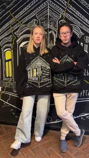 Двое людей в чёрных худи с принтами дома позируют на фоне графичного мурала; видно посадку изделия и как выглядит рисунок в интерьере.