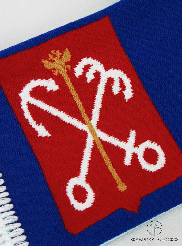 Крупный план вязаного герба Санкт‑Петербурга на красном фоне: перекрещенные якоря и скипетр, выполненные интарсией, хорошо видна структура нитей и плотность полотна.