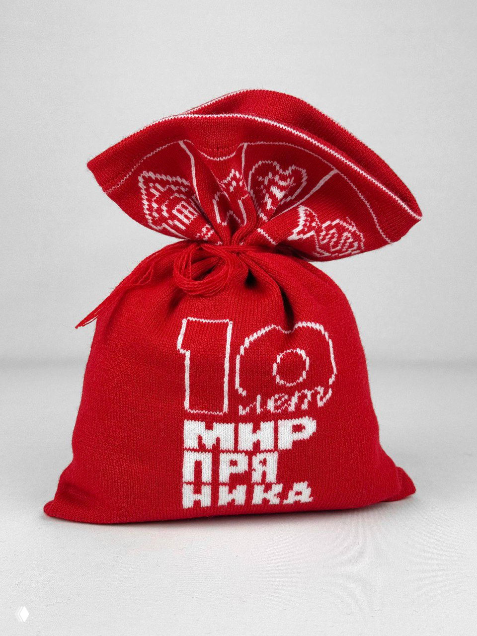 Вязаный мешочек со стороны: красное полотно и белая надпись «10 лет Мир пряника», изделие завязано декоративной петлей, студийный ракурс.