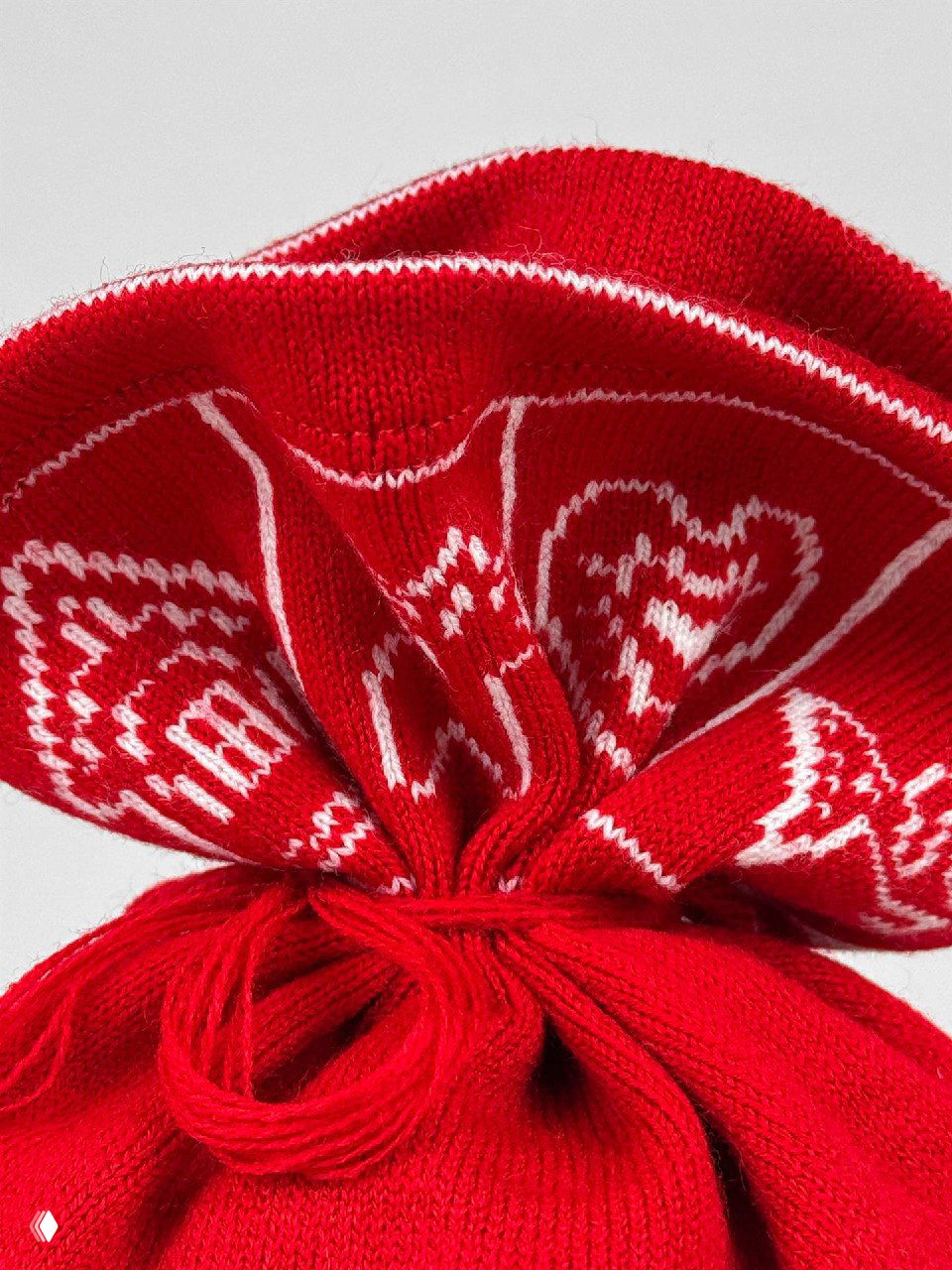 Крупный план верхней части мешочка: красная вязка с белыми узорами в виде сердечек и декоративной отделкой на фоне студии, текстура нитей видна.