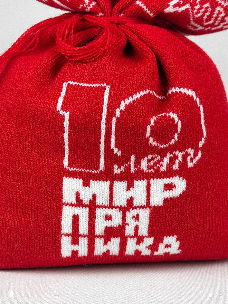 Детальный кадр узла и верхней части мешочка: красные нитки, аккуратно завязанный шнур и белые орнаменты на трикотаже, макрофотография изделия.