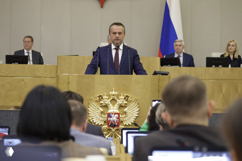 Выступающий у трибуны в зале Государственной думы РФ на фоне герба и флага; в кадре видны депутаты, столы и рабочие места в зале заседаний.