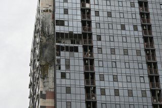 Фасад многоэтажного дома с пробоиной и следами разрушений после попадания дрона: выбитые окна и повреждённые панели.