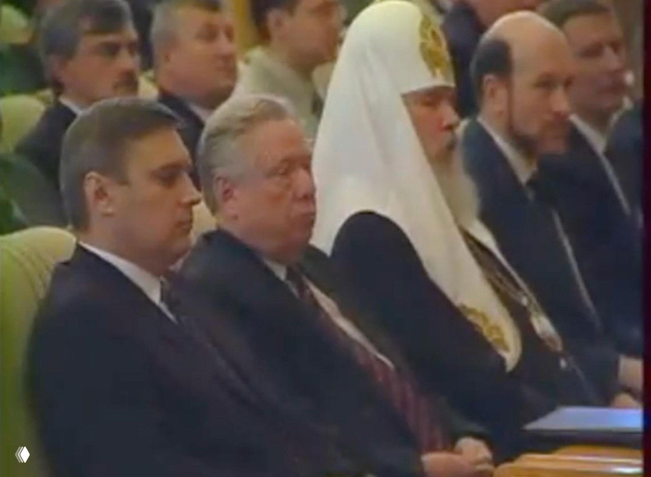 Архивное фото: первый ряд зала с представителями церковного и политического корпуса, мужчины в костюмах и церковной рясе, видна концентрация внимания.