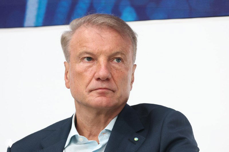 Крупный план мужчины в деловом костюме: серьёзное выражение лица, конференц-зал и зрительный зал на фоне, строгая деловая обстановка