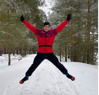 Бегун в красной куртке и черных штанах прыгает на заснеженной лесной тропе; видны зимние кроссовки и хвойные деревья.