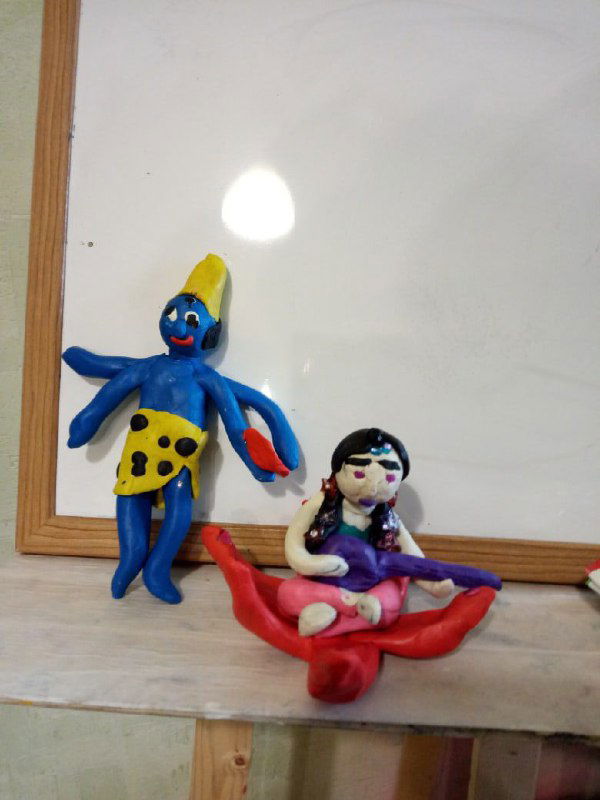 Небольшие пластилиновые фигурки, слепленные детьми: персонажи с простыми формами и яркими деталями, расположенные на фоне белой доски в мастерской.