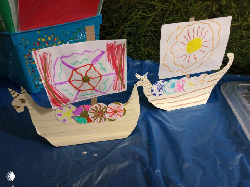 Бумажные или картонные макеты драккара, разрисованные детьми, выставлены на синем фоне для фотографирования работ.