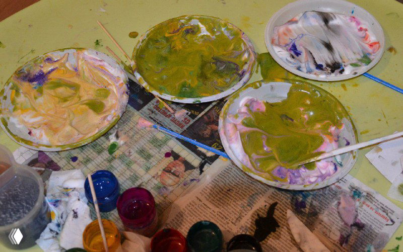 Тарелки с перемешанными красками и кистями на столе, видно хаотичное смешивание цветов как часть игрового процесса обучения детей.