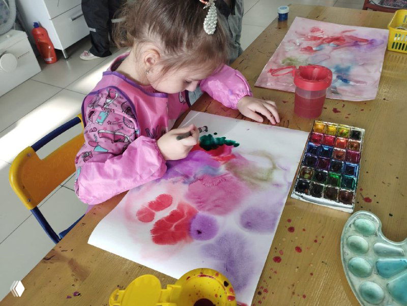 Ребёнок сосредоточенно рисует акварелью за столом; видны разноцветные пятна краски и рабочие материалы рядом, атмосфера эксперимента.