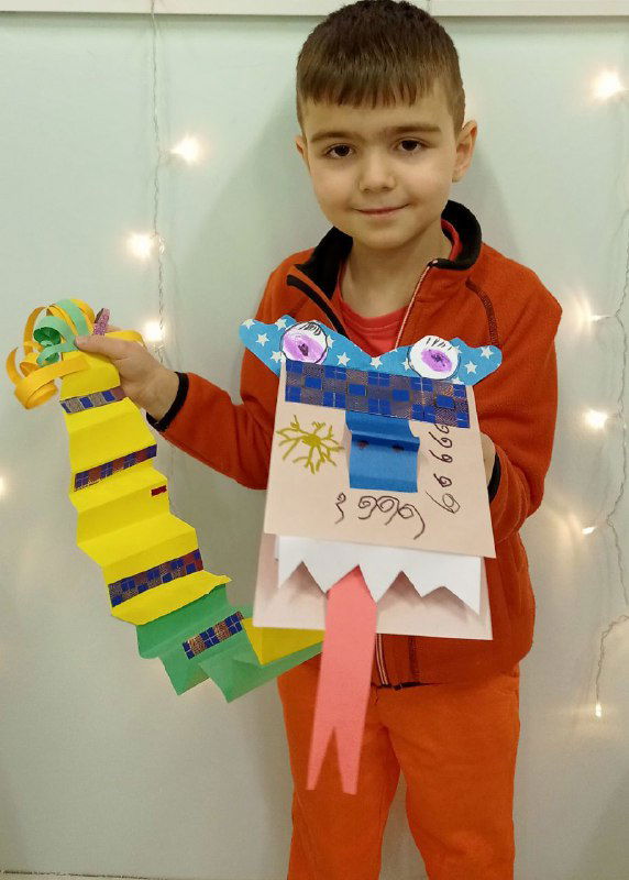 Мальчик держит жёлто-зелёного дракона с длинным хвостом из сложенной гармошкой бумаги; видно оформление головы и язык из полоски.