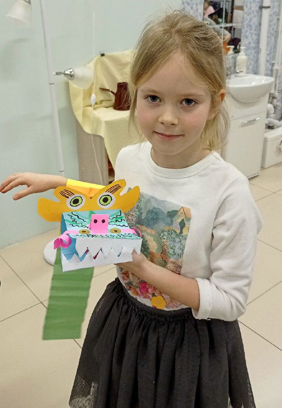 Девочка демонстрирует небольшого бумажного дракона с выразительными глазами и зубами; фон — класс с материалами для творчества.