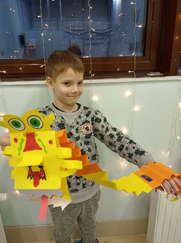 Мальчик держит крупного жёлтого бумажного дракона с развернутым хвостом; видно элементы декора и аккуратные складки гармошки.