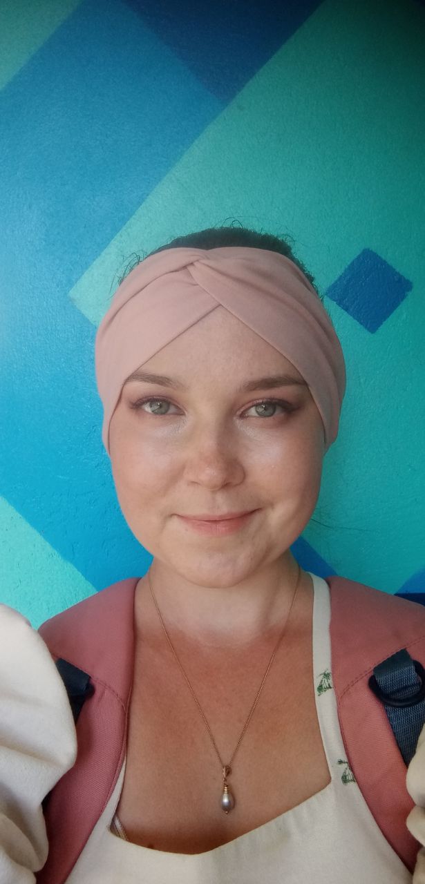 Селфи автора на фоне бирюзовой стены: мягкая улыбка, повязка на голове и ожерелье с подвеской, спокойное и светлое выражение лица.