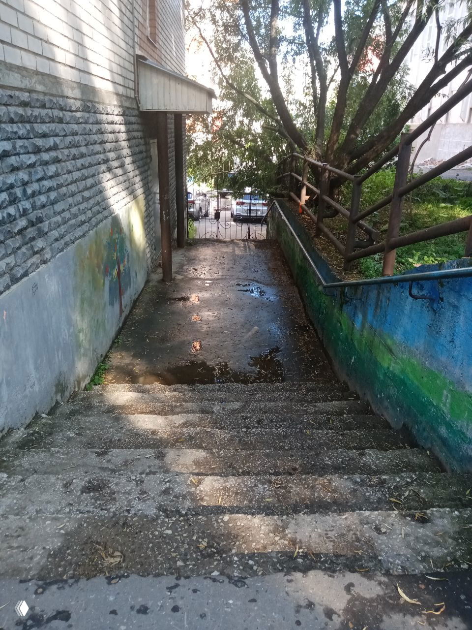 Спуск со ступенями между домами с мокрым асфальтом и железными перилами, тень деревьев и ограждение у выхода во двор.