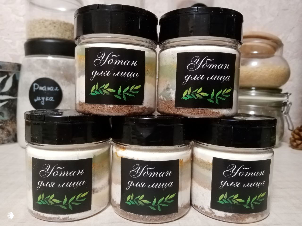 Пирамида из пяти прозрачных баночек с наклейками «Убтан для лица», в банках видны слои разных порошковых компонентов, на кухонном фоне банки и банки с ингредиентами.