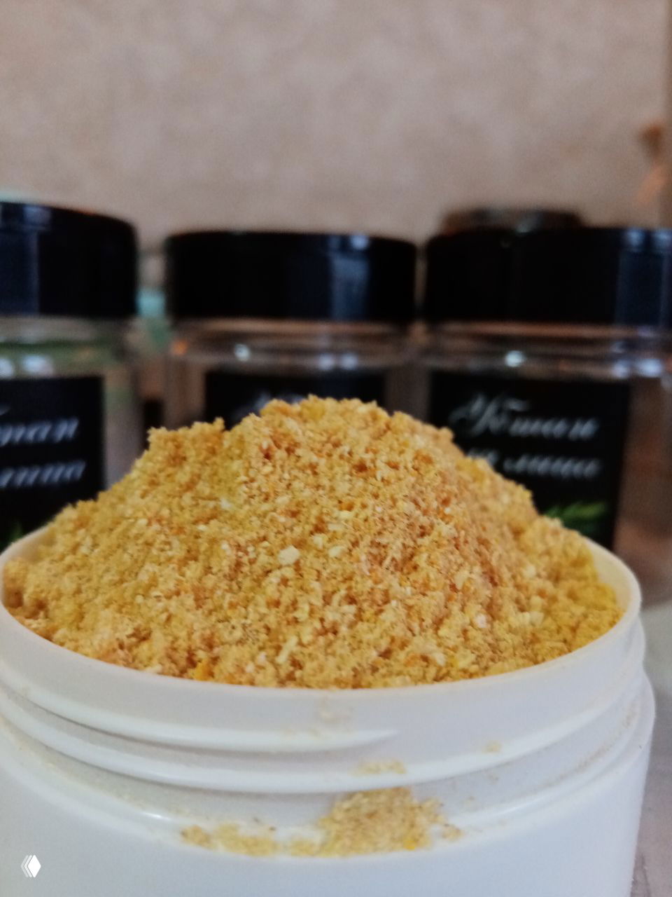 Открытая белая банка с горкой жёлто‑оранжевого порошка убтана на переднем плане; в размытом фоне видны подписанные баночки с наклейками.